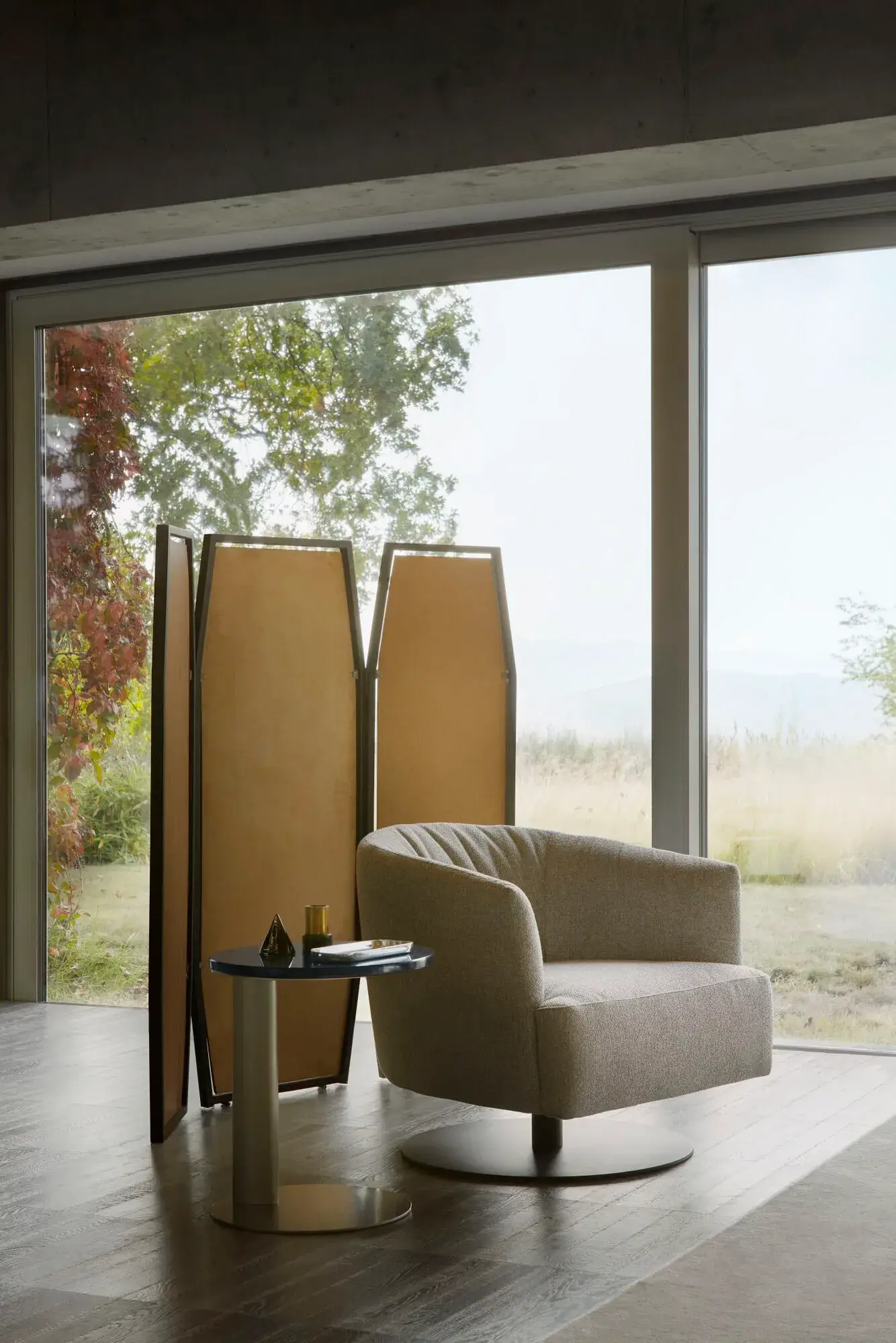 2b_NICOLINE_DIGA-SWIVEL_mkJHYrBfxO gray snug looking circular swivel arm chair on round black base in an empty room with large windows